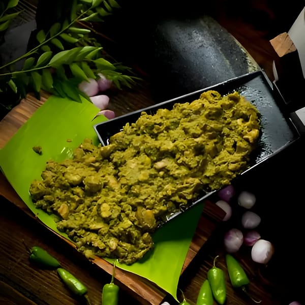 Green Indian curry on a black tray and banana leaf, with chilies, onions, and curry leaves.