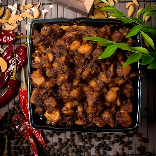 Indian chicken fry in a black dish, surrounded by red chilies, peppercorns, and green leaves.