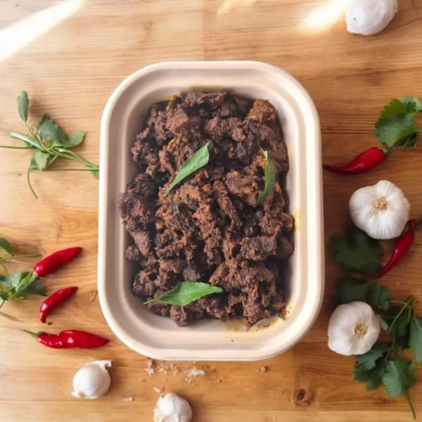 Kolapasi Indian Canteen's Indian beef fry in a container, surrounded by red chilies, garlic, and cilantro.